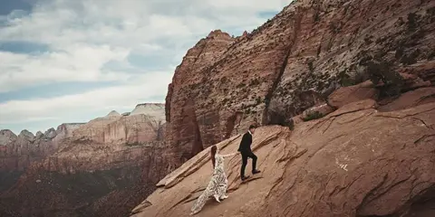 A bride and groom walking hand-in-hand on a rocky mountain slope with a vast canyon landscape in the background.