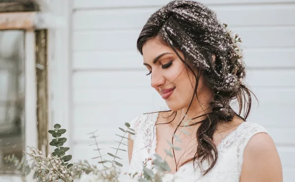 Bride in a white lace dress with a styled updo and floral hair accents, holding a bouquet in a snowy outdoor setting.