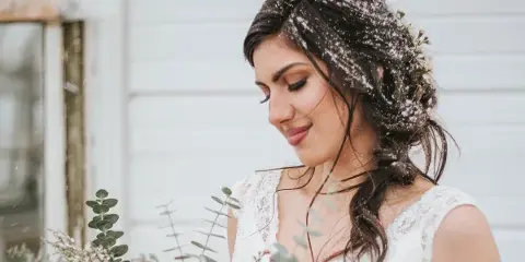Bride in a white lace dress with a styled updo and floral hair accents, holding a bouquet in a snowy outdoor setting.