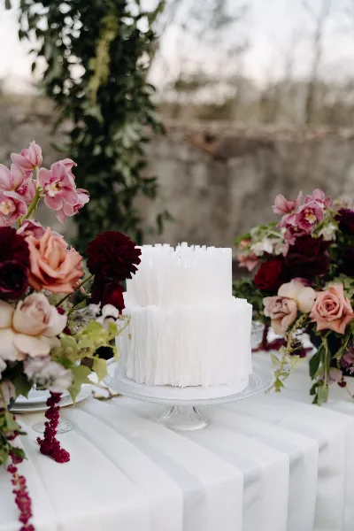 A white two-tier wedding cake with textured vertical detailing displayed on a glass cake stand, surrounded by pink and deep red floral arrangements on a white tablecloth.