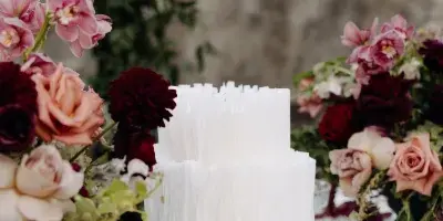 A white two-tier wedding cake with textured vertical detailing displayed on a glass cake stand, surrounded by pink and deep red floral arrangements on a white tablecloth.