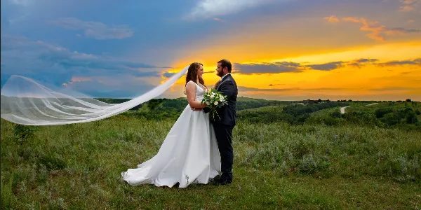 A bride and groom standing in a grassy field at sunset with the bride's veil blowing in the wind.