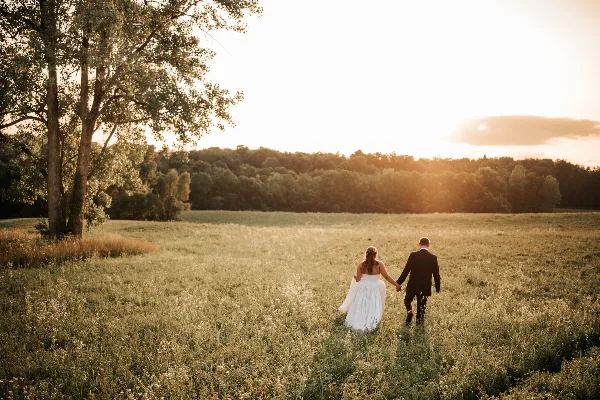 A bride and groom walking hand-in-hand through a grassy field at sunset with a forest in the background.
