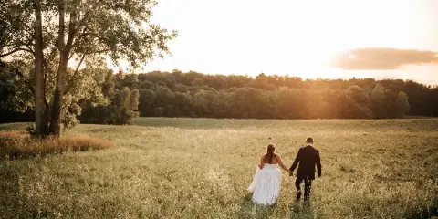 A bride and groom walking hand-in-hand through a grassy field at sunset with a forest in the background.