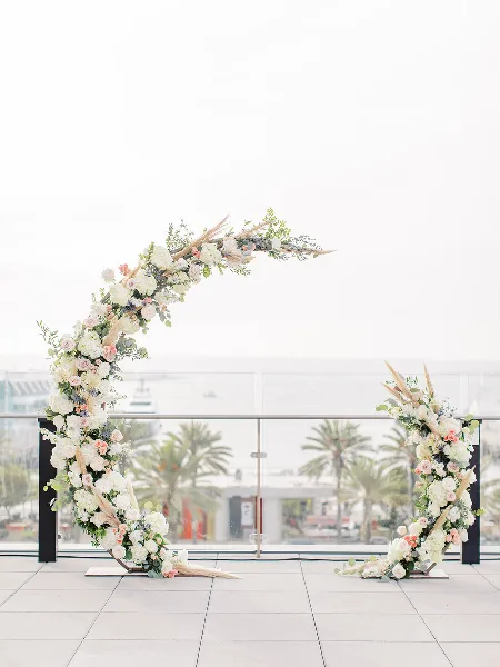 A modern, asymmetrical wedding arch decorated with white and pink flowers and greenery, set on a tiled terrace with a view of palm trees.
