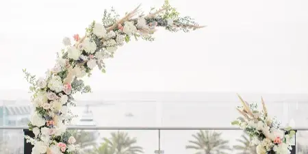 A modern, asymmetrical wedding arch decorated with white and pink flowers and greenery, set on a tiled terrace with a view of palm trees.