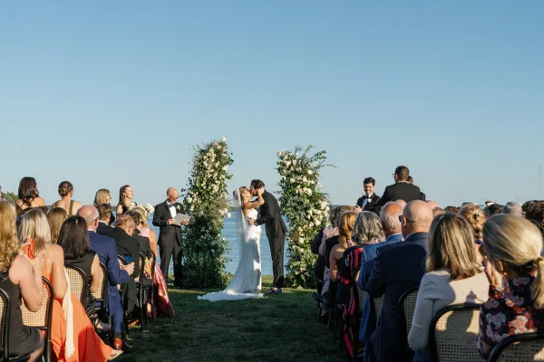 Outdoor wedding ceremony with a bride and groom kissing at the altar, surrounded by seated guests and floral arches.