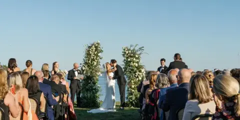 Outdoor wedding ceremony with a bride and groom kissing at the altar, surrounded by seated guests and floral arches.