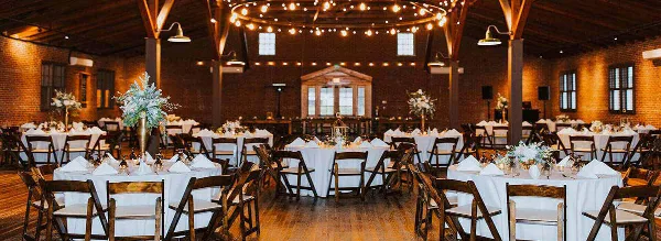 Wide shot of an industrial-style wedding reception hall with round tables, white linens, and string lights.