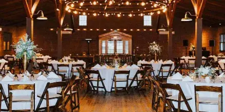 Wide shot of an industrial-style wedding reception hall with round tables, white linens, and string lights.