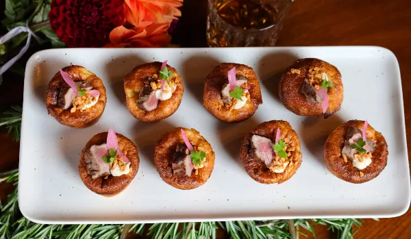 A white rectangular platter holding eight individual savory tarts or pastries, garnished with herbs and edible flowers, set on a wooden table with floral arrangements in the background.