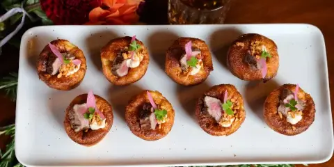 A white rectangular platter holding eight individual savory tarts or pastries, garnished with herbs and edible flowers, set on a wooden table with floral arrangements in the background.