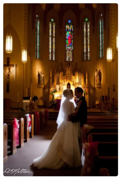 A bride and groom standing in the aisle of a church with stained glass windows and an altar in the background.