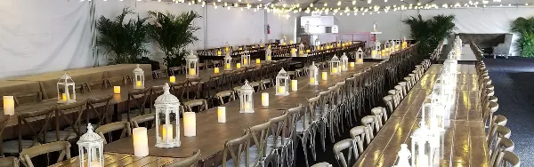 A long banquet table set with chairs, linens, and lanterns inside a white event tent.