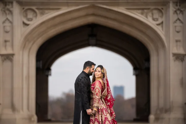 A bride and groom in traditional South Asian wedding attire standing together under a stone archway.