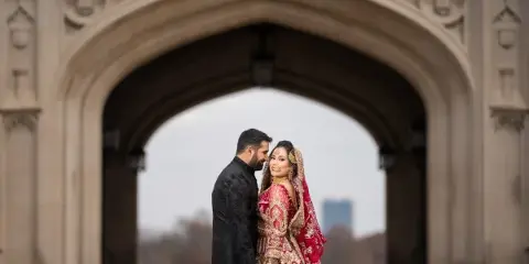 A bride and groom in traditional South Asian wedding attire standing together under a stone archway.