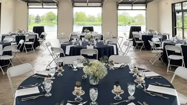 Interior view of a wedding venue ballroom with round tables set for a reception and large glass doors overlooking a body of water.