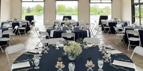 Interior view of a wedding venue ballroom with round tables set for a reception and large glass doors overlooking a body of water.