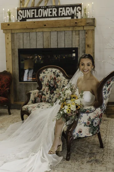 A bride in a white wedding dress sits in a floral armchair holding a bouquet, with a rustic wooden fireplace and a 'Sunflower Farms' sign in the background.