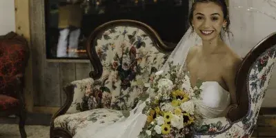 A bride in a white wedding dress sits in a floral armchair holding a bouquet, with a rustic wooden fireplace and a 'Sunflower Farms' sign in the background.