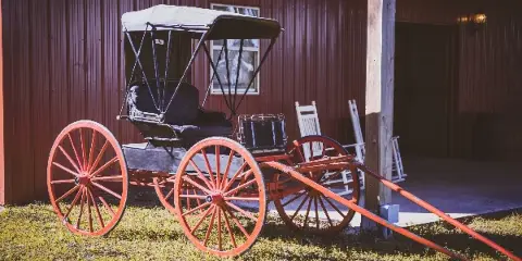 A red vintage carriage parked on a grassy area in front of a rustic red building.