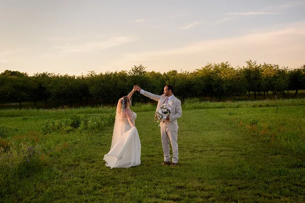 A bride and groom dancing in a grassy field at sunset with an orchard in the background.