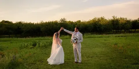 A bride and groom dancing in a grassy field at sunset with an orchard in the background.