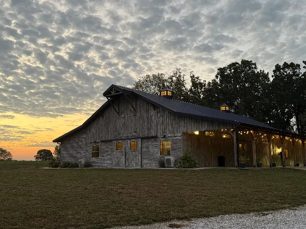 Exterior view of a large wooden event barn with a covered porch and string lights at dusk.
