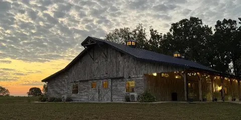 Exterior view of a large wooden event barn with a covered porch and string lights at dusk.