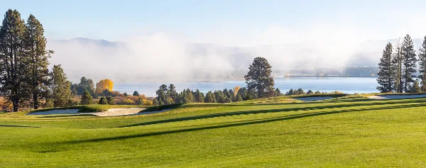 A scenic view of the Tamarack Resort golf course with manicured greens and sand traps overlooking a lake and misty mountains.