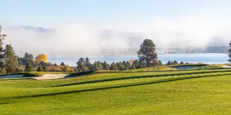 A scenic view of the Tamarack Resort golf course with manicured greens and sand traps overlooking a lake and misty mountains.