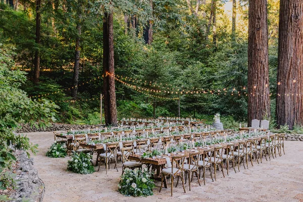 An outdoor wedding reception setup featuring long wooden tables, white chairs, floral arrangements, and string lights in a forest clearing.