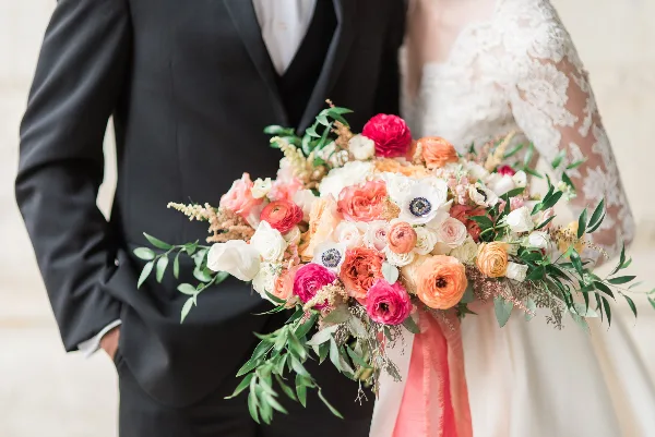 A close-up of a colorful wedding bouquet featuring pink, orange, and white flowers held by a bride and groom.