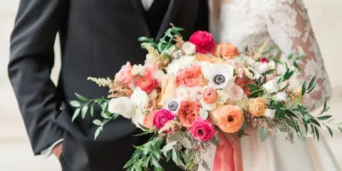 A close-up of a colorful wedding bouquet featuring pink, orange, and white flowers held by a bride and groom.
