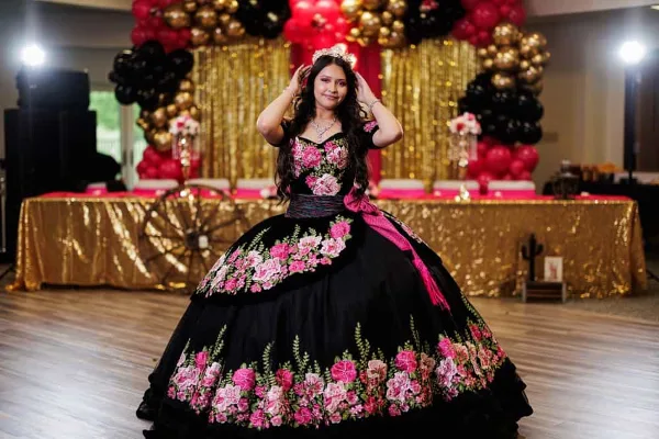 A young woman in a formal black and pink floral dress poses on a dance floor in front of a decorated backdrop with balloons and gold drapery.