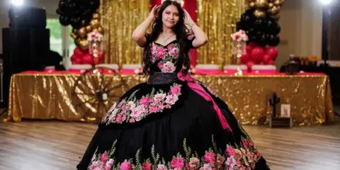 A young woman in a formal black and pink floral dress poses on a dance floor in front of a decorated backdrop with balloons and gold drapery.