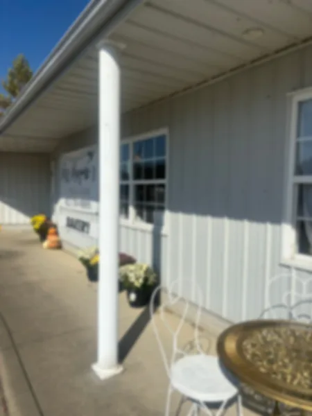 Exterior view of Miss Mayme's Bakery storefront with white siding, a porch column, potted flowers, and outdoor seating.