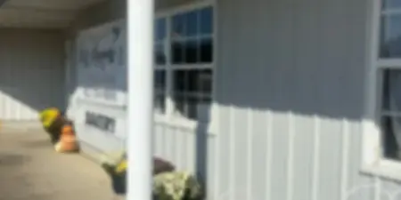 Exterior view of Miss Mayme's Bakery storefront with white siding, a porch column, potted flowers, and outdoor seating.