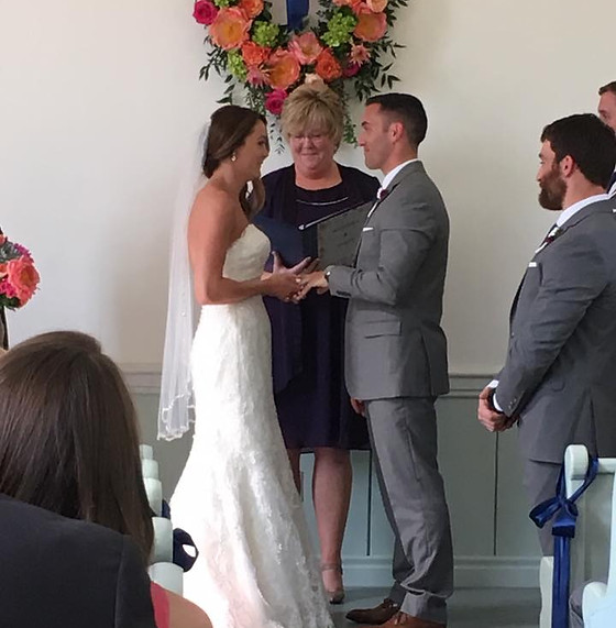 A wedding officiant stands between a bride and groom during a ceremony, with guests seated in the foreground.