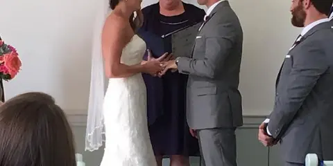 A wedding officiant stands between a bride and groom during a ceremony, with guests seated in the foreground.