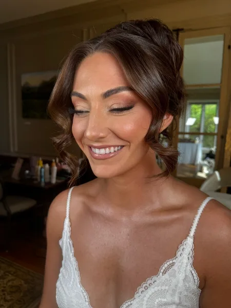 A smiling bride in a white lace dress with an updo hairstyle and professional makeup.