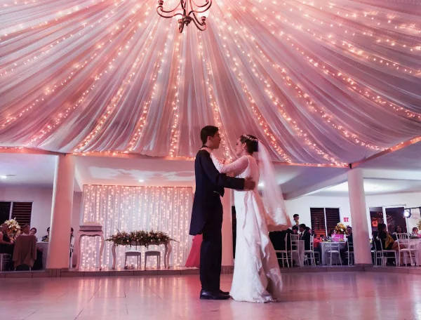 A bride and groom dancing at a wedding reception under a ceiling decorated with draping fabric and string lights.