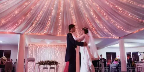 A bride and groom dancing at a wedding reception under a ceiling decorated with draping fabric and string lights.