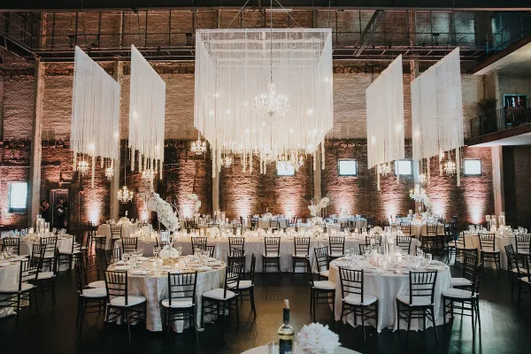 A wedding reception hall featuring round tables, white linens, and large hanging white drapery installations with chandeliers.
