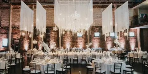 A wedding reception hall featuring round tables, white linens, and large hanging white drapery installations with chandeliers.
