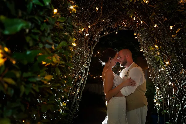 A bride and groom embracing under a leafy archway decorated with string lights at night.