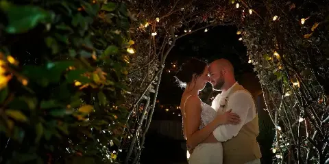 A bride and groom embracing under a leafy archway decorated with string lights at night.
