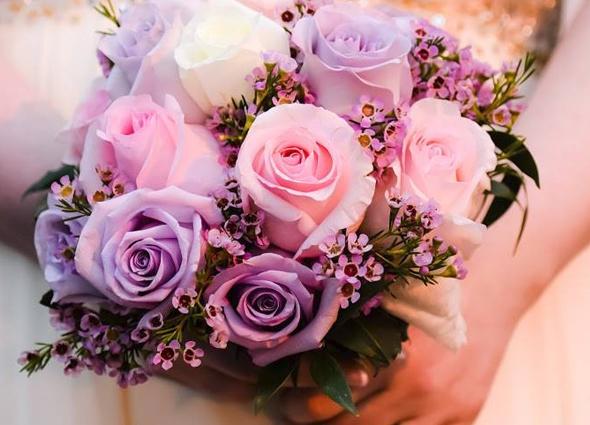 A hand holding a bridal bouquet of pink, white, and lavender roses with small purple filler flowers.