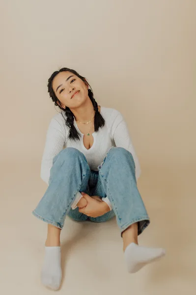 A young woman with braided hair sitting on the floor in a white top and jeans, photographed in a studio setting.