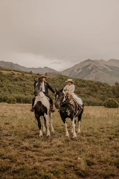 A bride and groom in western-style wedding attire riding horses side-by-side in a grassy field with mountains in the background.